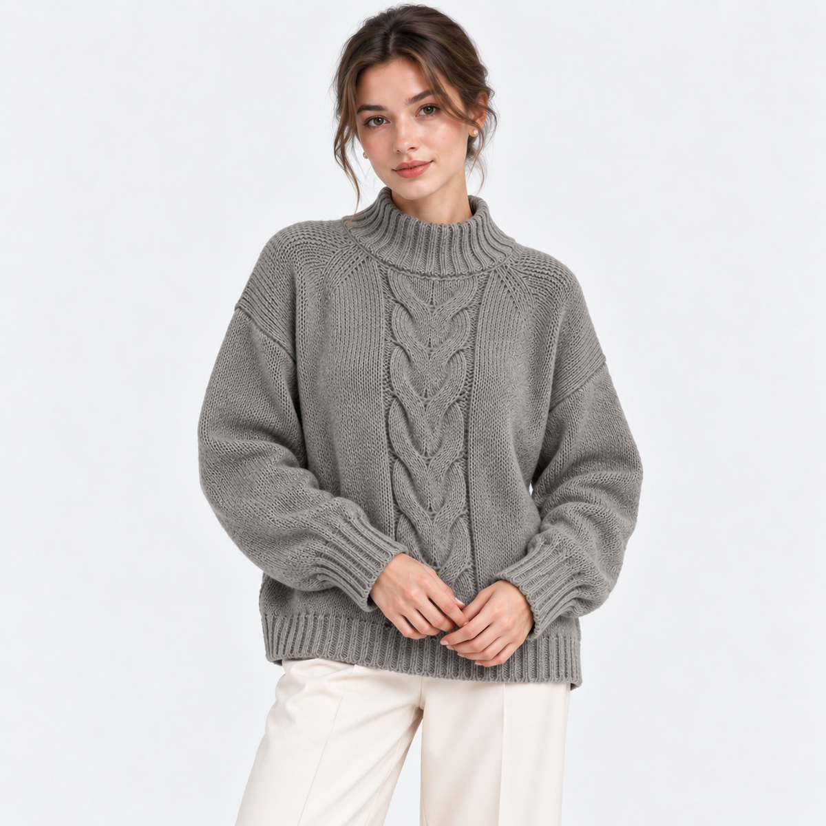 A woman wearing a gray cable-knit mock neck sweater and cream pants, standing against a plain white studio background.