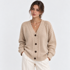 A woman wearing a beige cable-knit cardigan sweater with dark buttons and cream pants, standing against a plain light gray studio background.