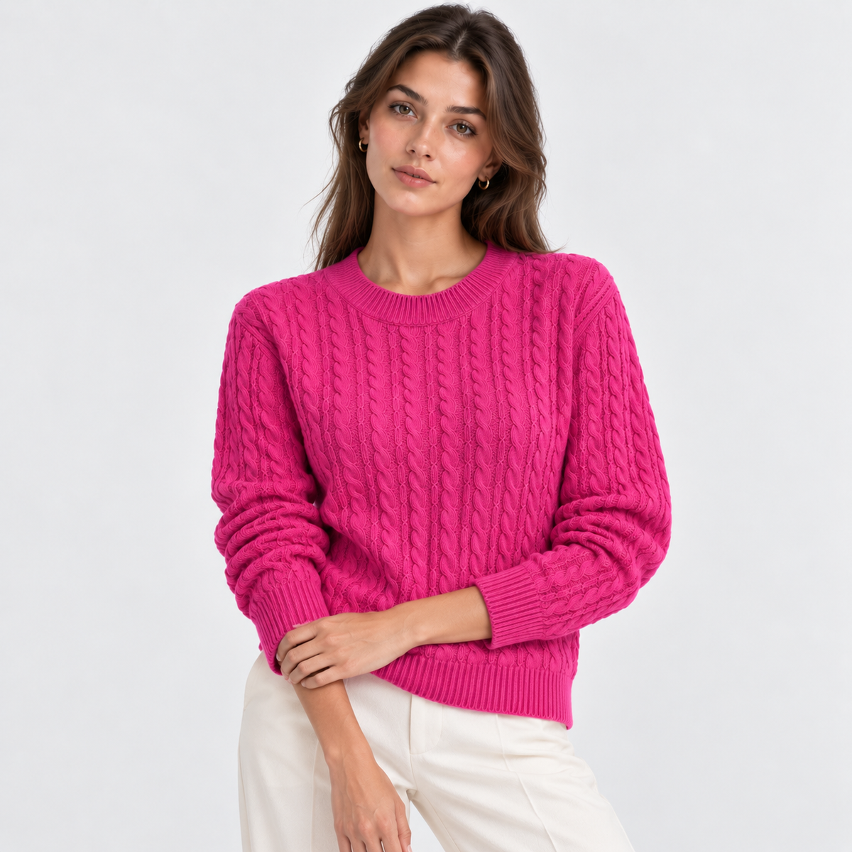 A woman wearing a bright pink cable-knit crewneck sweater and white pants, standing against a light gray studio background.