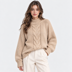 A woman wearing a beige cable-knit mock neck sweater and cream-colored trousers, standing against a solid light gray background.