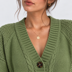 A close-up of a woman wearing an olive green cable-knit cardigan sweater with a brown button, styled with a delicate gold disc necklace and gold hoop earrings against a light gray background.