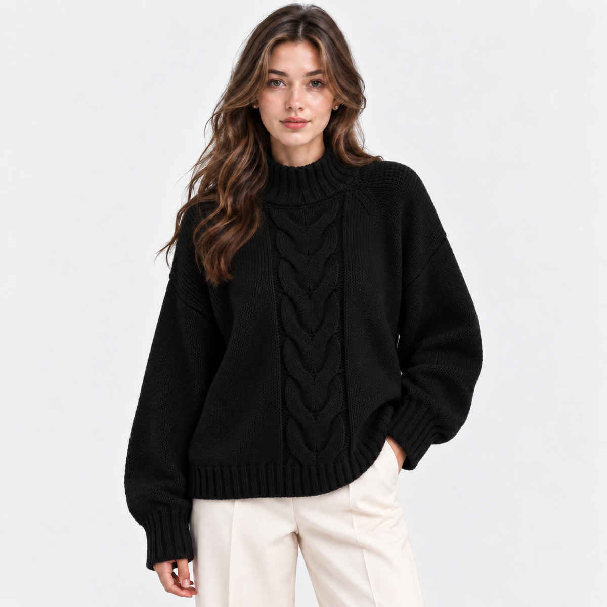 A woman wearing a black cable-knit mock-neck sweater and cream-colored pants, standing against a plain light gray studio background.