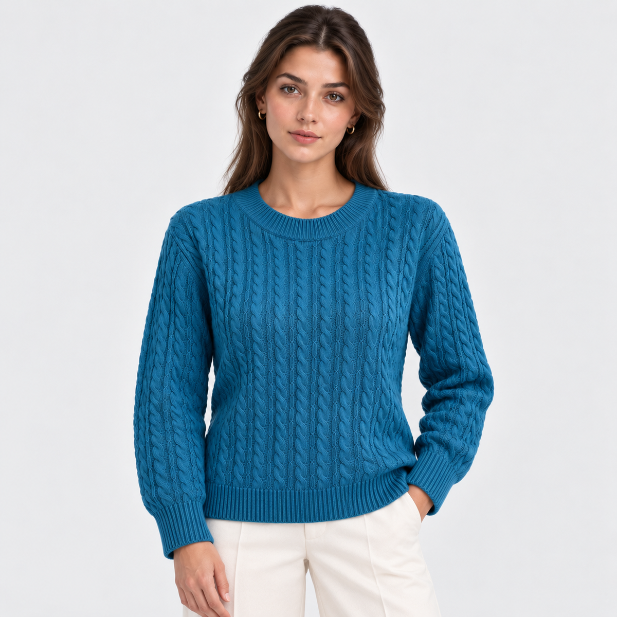 A woman modeling a teal blue cable-knit crewneck sweater with white pants, photographed against a clean light gray studio background.