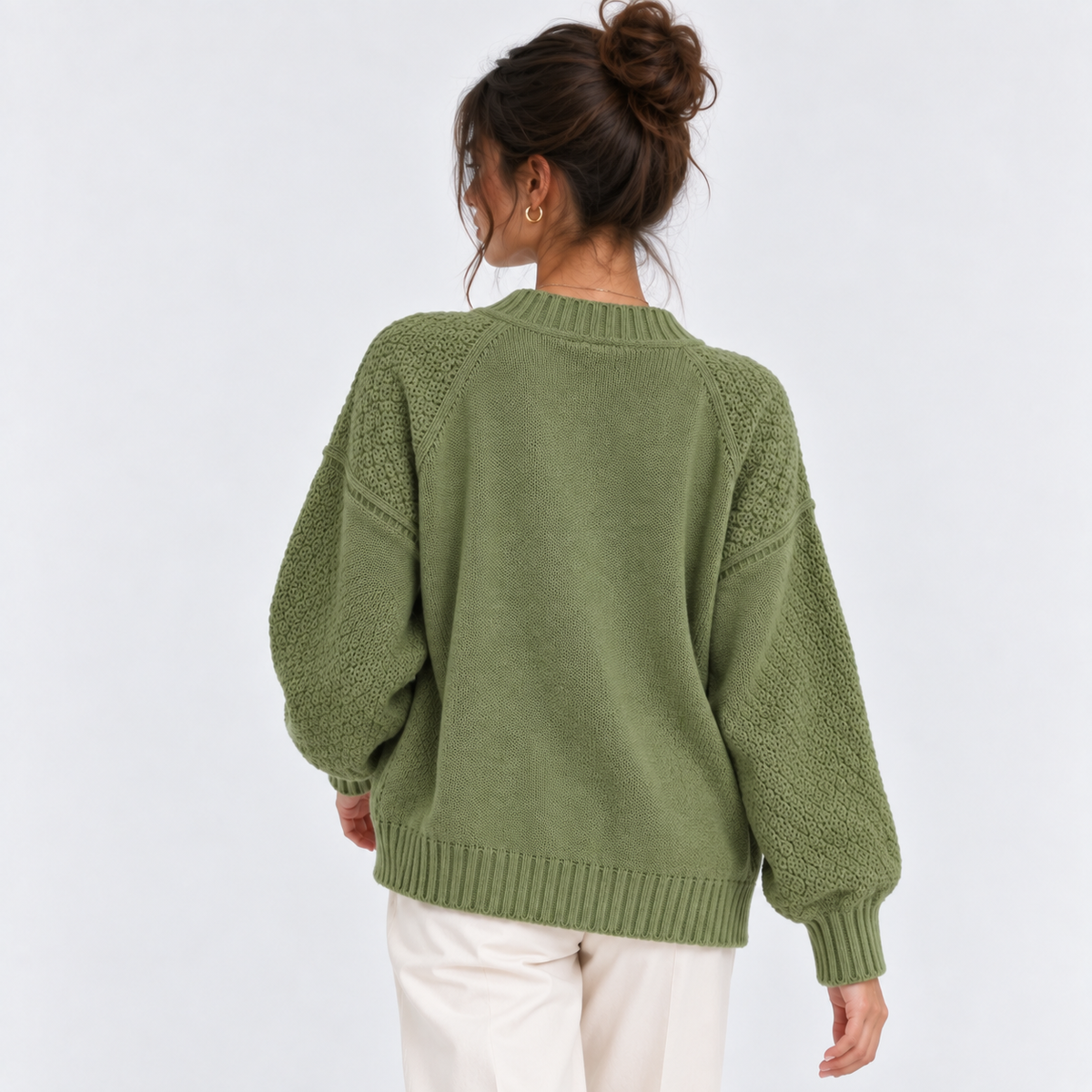 A woman viewed from the back wearing an olive green knit sweater with textured, chunky sleeves and cream pants, set against a plain light gray studio background.
