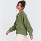 A woman wearing an olive green cable-knit cardigan sweater with dark buttons, paired with white pants, photographed against a clean white studio background.