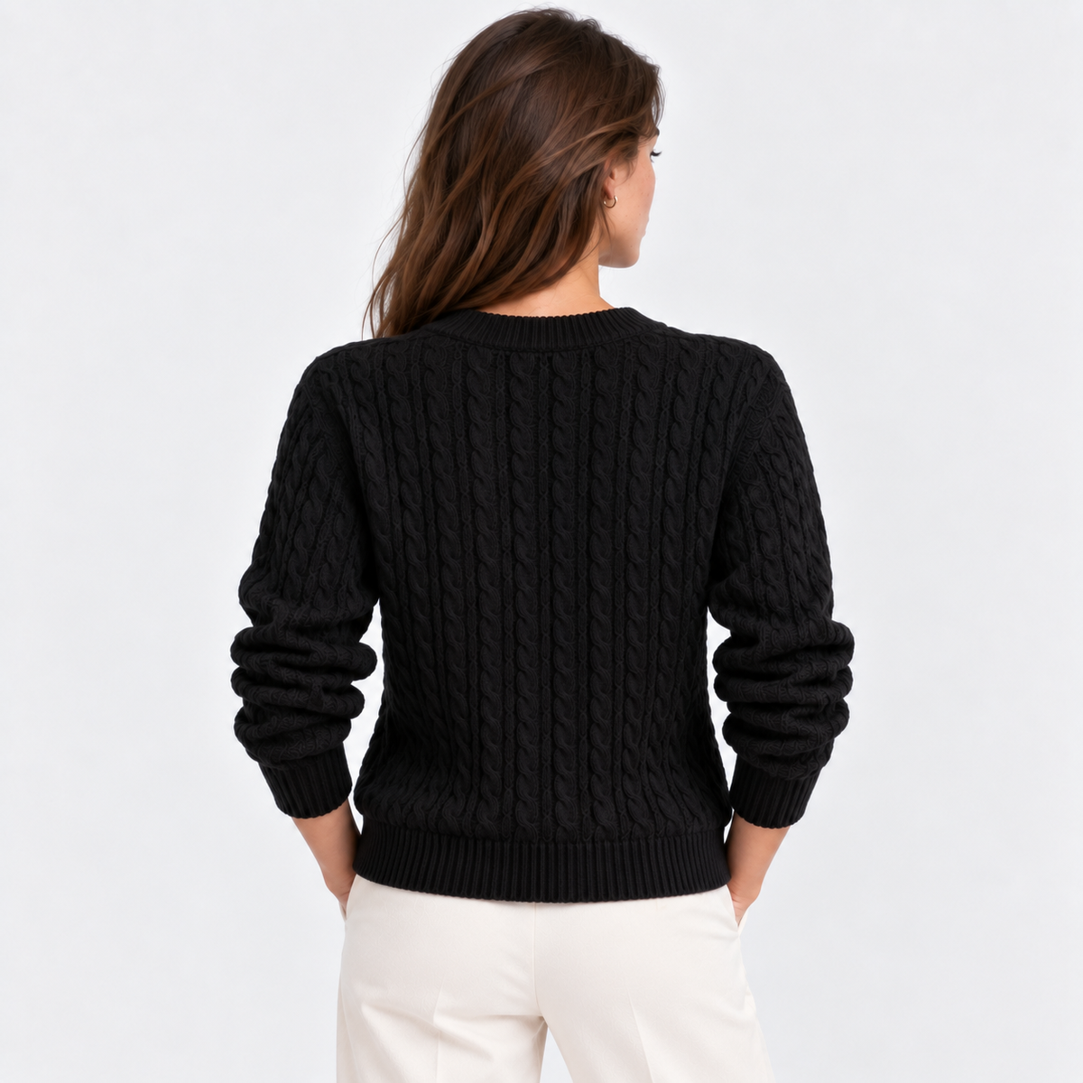 A woman viewed from the back wearing a black cable-knit sweater and off-white pants against a plain light gray studio background.