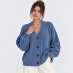 A woman wearing a blue cable-knit cardigan sweater with brown buttons, paired with cream pants, against a plain light gray studio background.