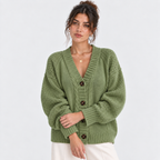 A woman wearing an oversized sage green knit cardigan with brown buttons, paired with white pants, against a plain light gray studio background.