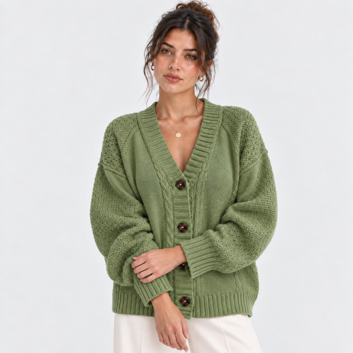 A woman wearing an oversized sage green knit cardigan with brown buttons, paired with white pants, against a plain light gray studio background.