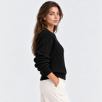 A woman wearing a black cable-knit sweater and cream-colored trousers, shown from a side profile against a plain light gray studio background.