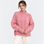 A woman wearing a pink cable-knit mock neck sweater and cream pants, standing against a plain white studio background.