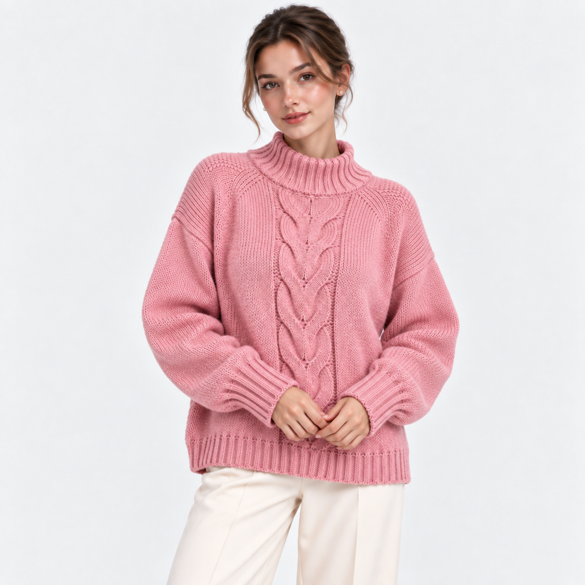 A woman wearing a pink cable-knit mock neck sweater and cream pants, standing against a plain white studio background.