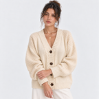 A woman wearing a cream-colored cable-knit cardigan sweater with dark brown buttons, paired with white pants, against a light gray studio background.