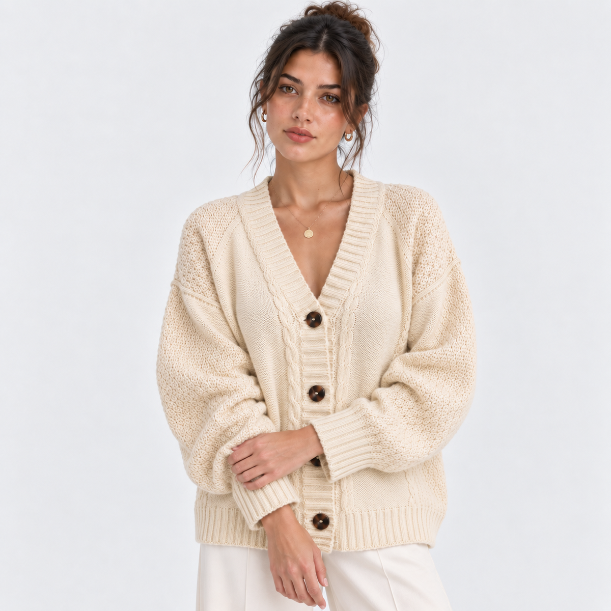 A woman wearing a cream-colored cable-knit cardigan sweater with dark brown buttons, paired with white pants, against a light gray studio background.