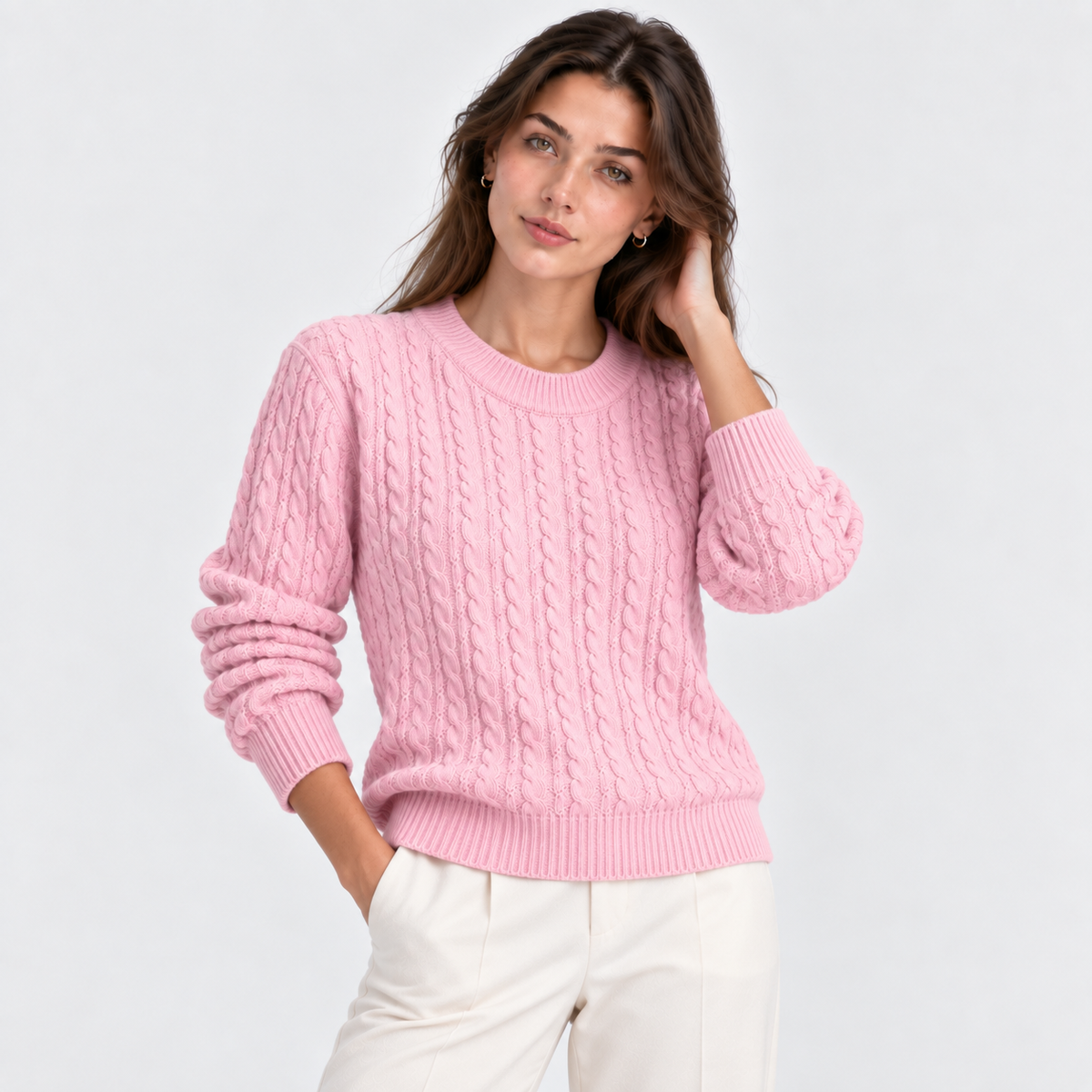 A woman wearing a light pink cable-knit crewneck sweater and white trousers, posing against a light gray studio background.