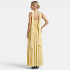 Back view of a woman wearing a pale yellow maxi dress with a lace-up back and flowing scarf detail against a light gray background.