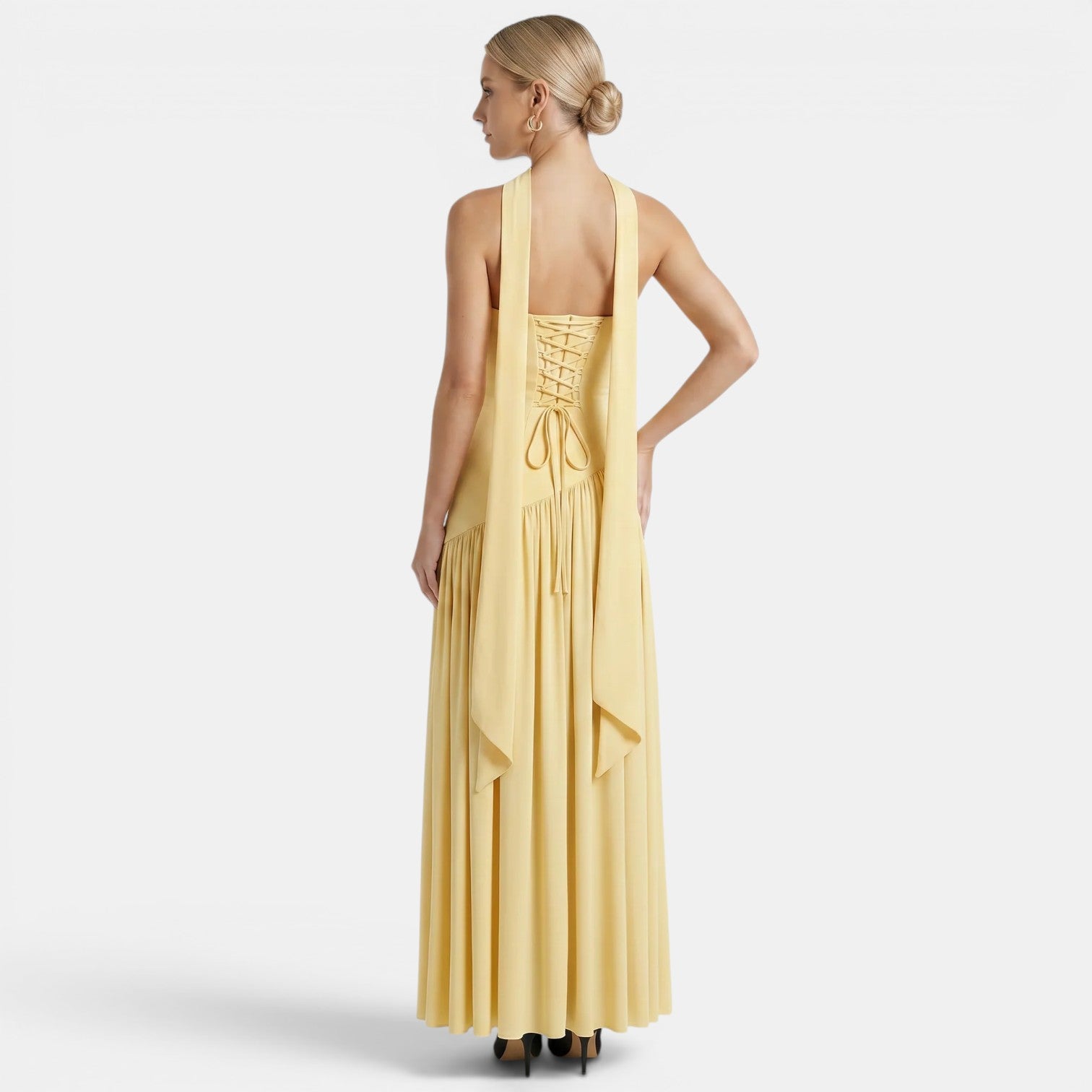 Back view of a woman wearing a pale yellow maxi dress with a lace-up back and flowing scarf detail against a light gray background.