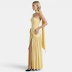 Side view of a woman wearing a pale yellow strapless maxi dress with a scarf neckline and thigh-high slit against a light gray background.
