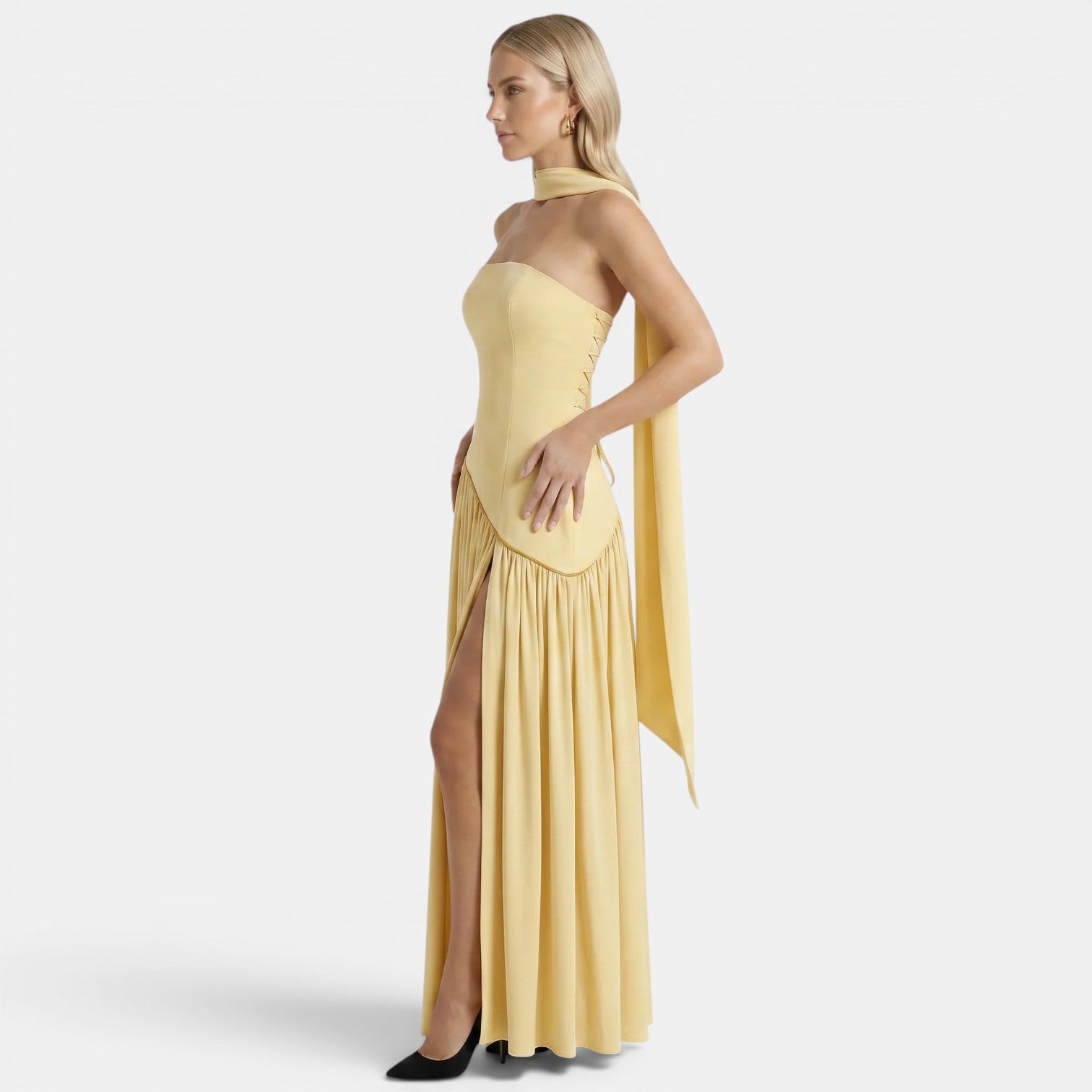 Side view of a woman wearing a pale yellow strapless maxi dress with a scarf neckline and thigh-high slit against a light gray background.