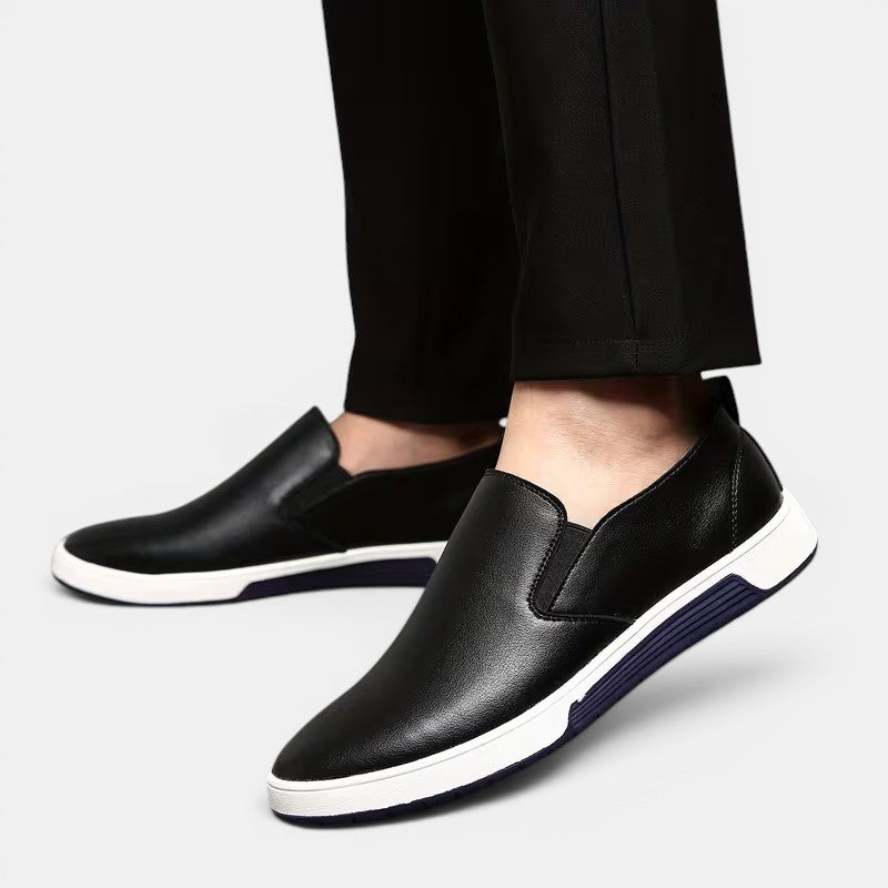 Black slip-on shoes with white soles worn with black pants on a light gray background