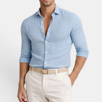 Man wearing a light blue button-up shirt and white pants on a plain background