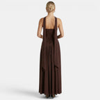 Back view of a woman wearing a dark brown maxi dress with a lace-up back and flowing scarf detail against a light gray background.