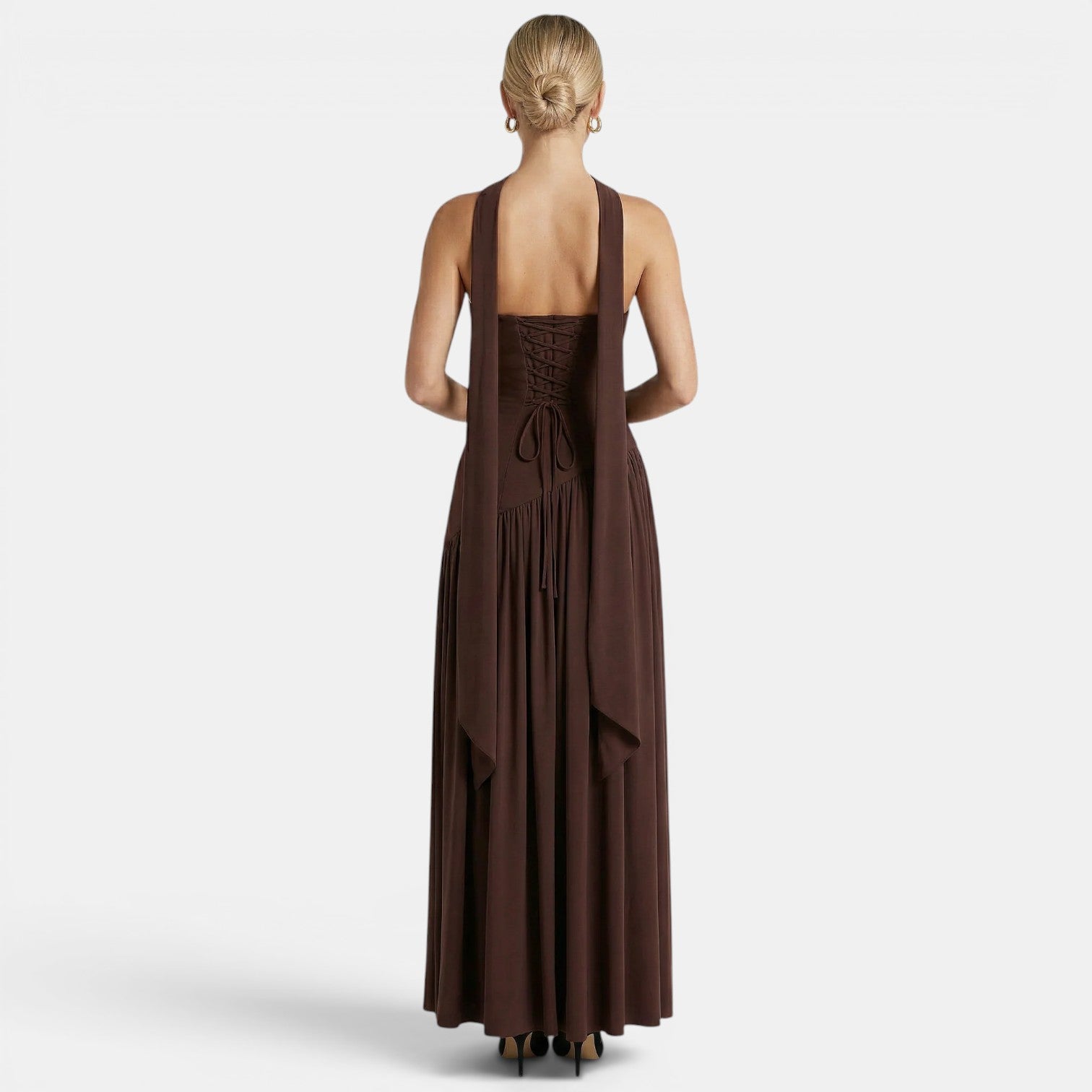 Back view of a woman wearing a dark brown maxi dress with a lace-up back and flowing scarf detail against a light gray background.