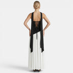 Back view of a woman wearing a black-and-white maxi dress with a lace-up back and flowing scarf detail against a light gray background.
