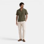 Man wearing an olive green textured knit polo shirt with cream trousers and brown loafers, standing against a light gray background.