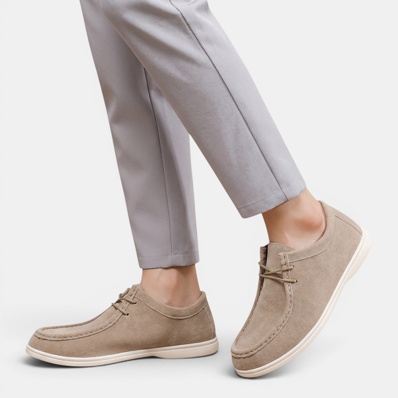 Person wearing beige suede lace-up shoes with white soles and light gray trousers, shown in a side walking pose.