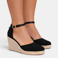 Black wedge sandals with espadrille sole worn by a person on a white background
